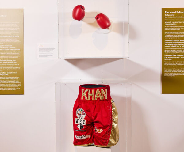 Red and Gold boxing shorts saying 'KHAN' in a clear box, and above the shorts are red boxing gloves in a clear box.