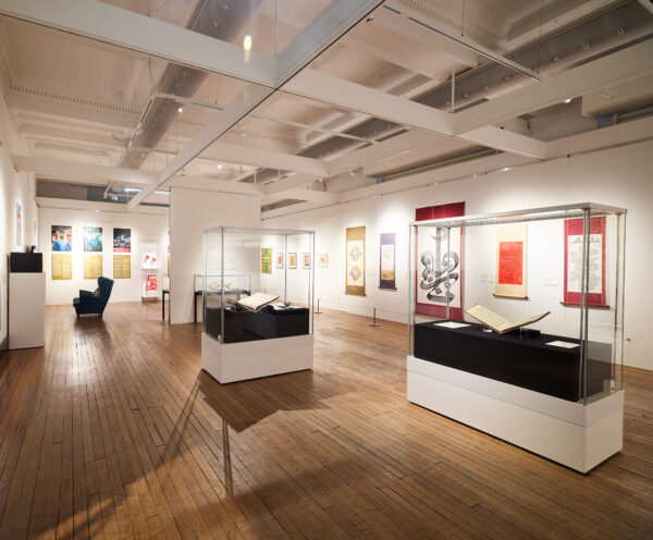 bright white exhibition room with multiple calligraphy art pieces hung on the walls and clear boxes in the centre of the room containing Quran books on display