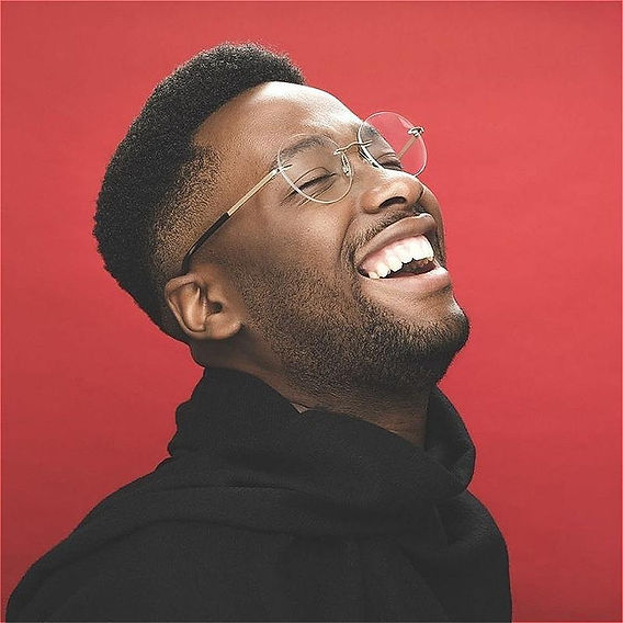 A black man with glasses laughs, he is wearing a black top and the background is red.