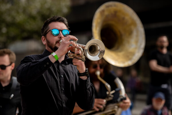 A man plays a trumpet, he is wearing shades and a black shirt.