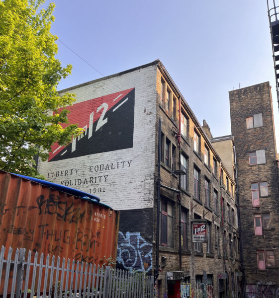 A large old mill towers above, with a black and red sign that reads '1 in 12' in white text. The sky is blue and there are green leaves on the tree to the left.