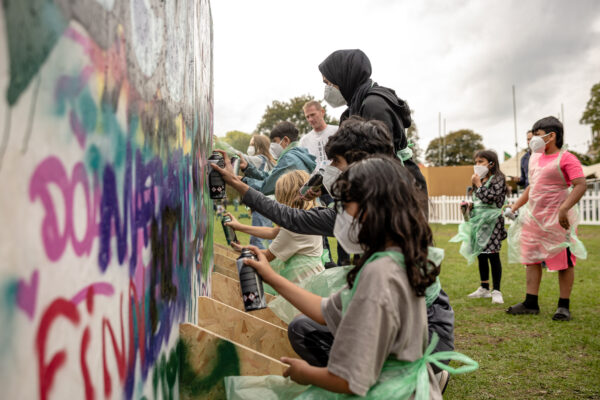 Lots of young people spray painting and painting on a collaborative wall mural