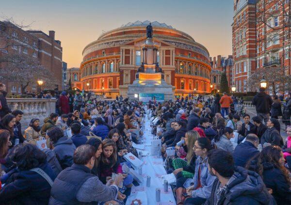 Lots of people sat on the floor in lines along long tables outside in a street, in front of a round building and statue. The people have paper bags and cans in front of them.