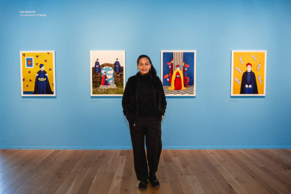 Full body image of lady (Aida Muluneh) in all black, posing in front of a blue wall with 4 images in a line across the wall