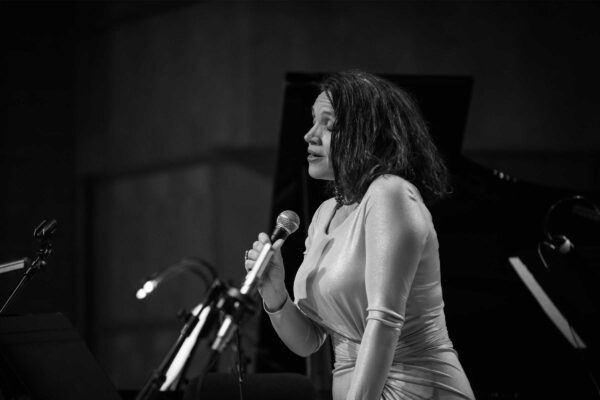 A black and white photograph of a woman in a silky white dress and elbow length gloves singing into a microphone