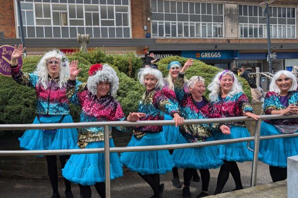 A group od women. in silver wigs and pink and blue outfits dance in a town square