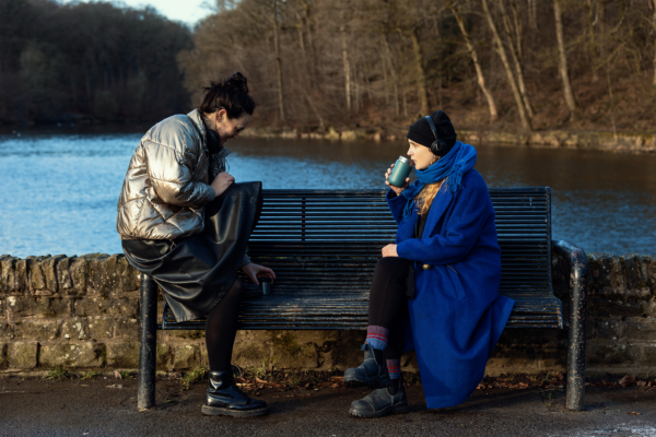 two women sat facing eachother on a bench, one is wearing headphones and drinking coffee