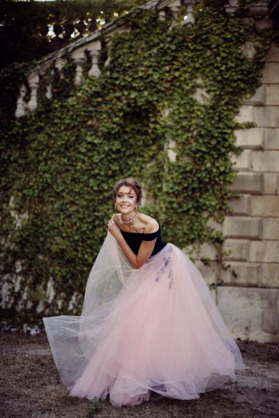 A woman poses before an ivy covered stone wall - she is wearing a dress made up of a pink tulle skirt and a black, off the shoulder top. He hair is pinned up and she is smiling.