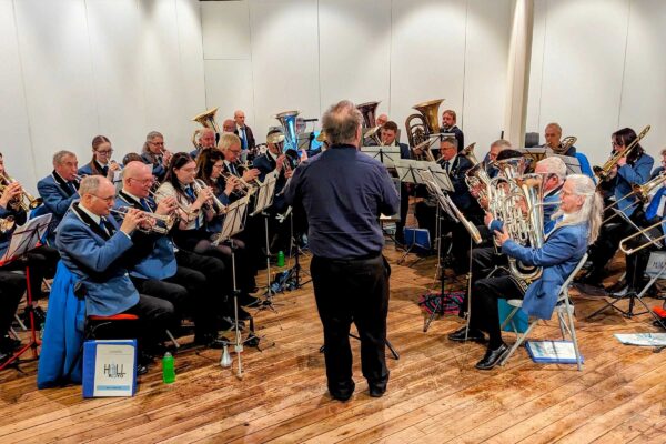 A brass band sat playing in a hall being conducted by a man who has his back to the camera