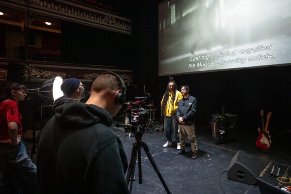 Two men from Asain Dub Foundation in front of the camera with drums and guitar at either side of them. Man with headphones and two other individuals stood behind the camera