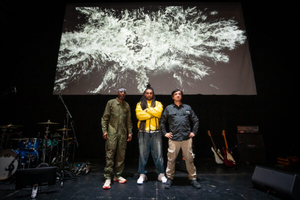 Image of three men, on stage posing infront of drums and guitairs