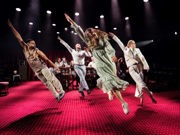 Four dancers leaping in syncronisation with one arm up in the air.