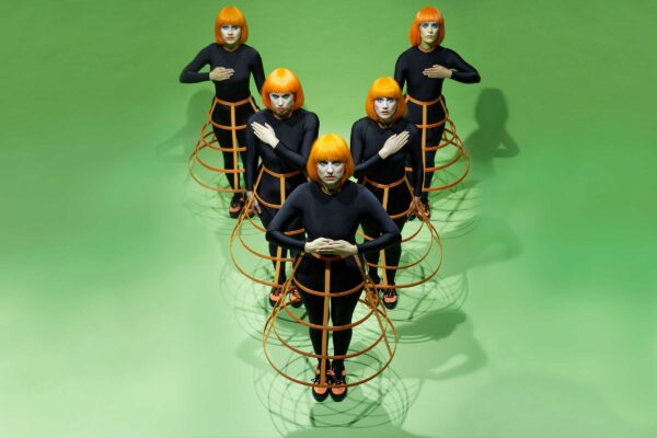 Five performers wearing orange wigs stand in a V shape, against a green background.