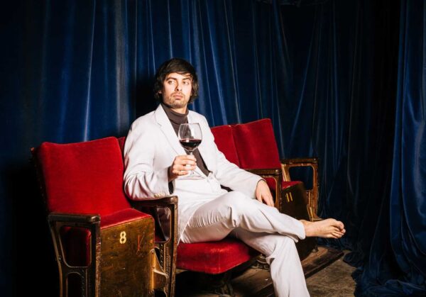 Man in a light grey suit sat on red theatre seats, holding a glass of red wine.