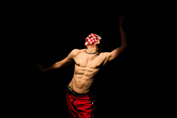 black background with a male int he spotlight, with his torso on show, red shorts and white and red checkered face paint on