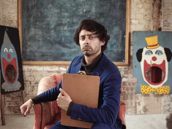 Man pulling a serious face to the camra, sat on a rustic red chair, holding a clipboard in front of him. In the background either side of him are two large drawings of clowns.