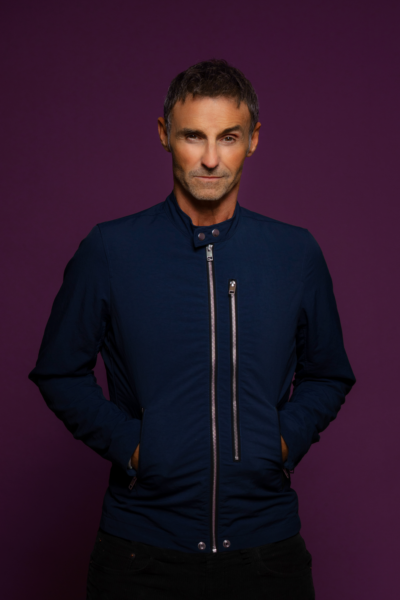 Headshot of Marti Pellow - a man looks to the camera, he has short, dark hair and is wearing a dark top with silver zips. The background is a dark reddish purple.