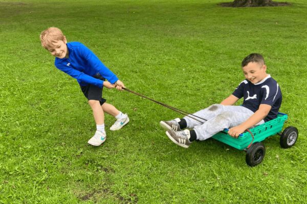 A young boy pulls another young boy who is sat in a makeshift cart across some grass