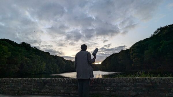 A person stands by a lake, holding a large microphone.