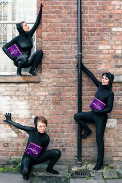 Three people wearing all black suits, holding boxes of Milk Tray.