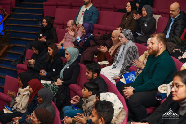 People sat in rows in an auditorium, they are clapping and looking engaged at the stage.