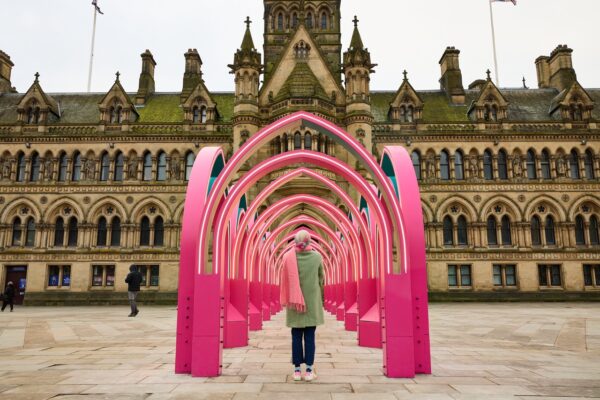 The Ramadan Pavilion, comprises of several arches that take inspiration from Bradford's City Hall. The pavilion is a bright pink and sits in front of the City Hall, and there is a woman stood within the arches of the pavilion.