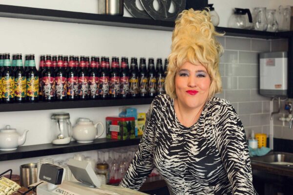 A woman with blonde beehive hair and a zebra print top stands behind a bar stacked with beer bottles