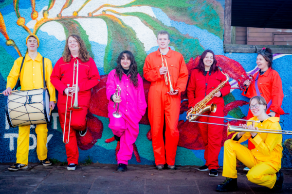 A group of people in brightly coloured jumpsuits pose with their instruments in front of a botanical mural