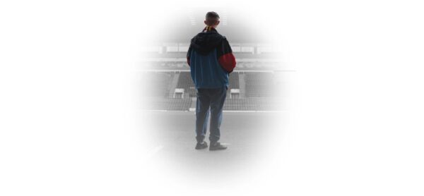 A man stands looking away from the camera - he is wearing a tracksuit stop and trousers. He's looking at a football stadium from the pitch, and is surrounded by white.