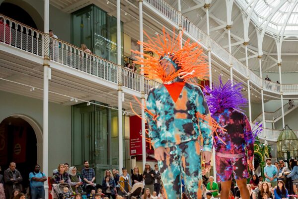 Two people dance back to back in brightly coloured outfits to a crowd of people in a heritage building