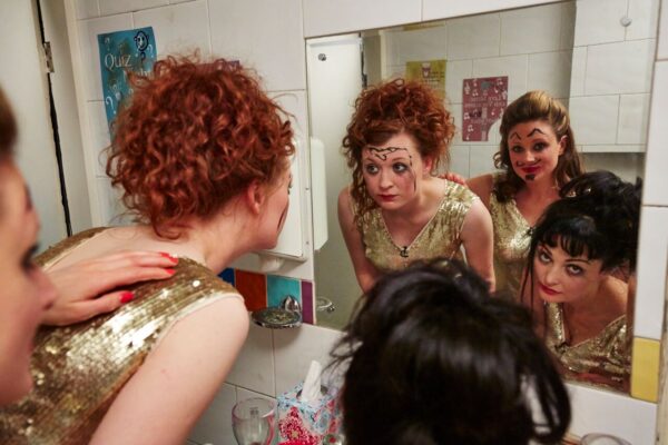 Three women look into a mirror. They wear gold dresses and have drawings on their faces. One of them has a penis drawn on her forehead.