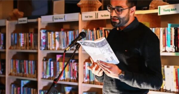 A photo of a man in glasses stood in front of shelves of books, speaking to a room of people