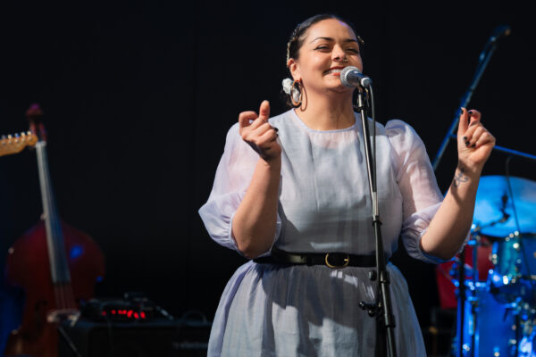 A white woman with dark hair sings into a microphone - she is wearing a blue dress with a black belt