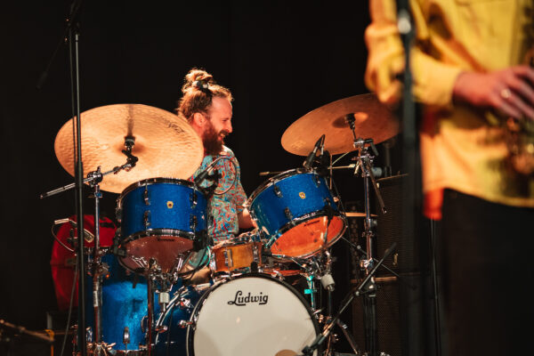 A white man with his hair in a bun plays the drums.