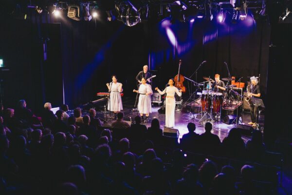Three women in white outfits sing to an audience - there is a full band behind them.