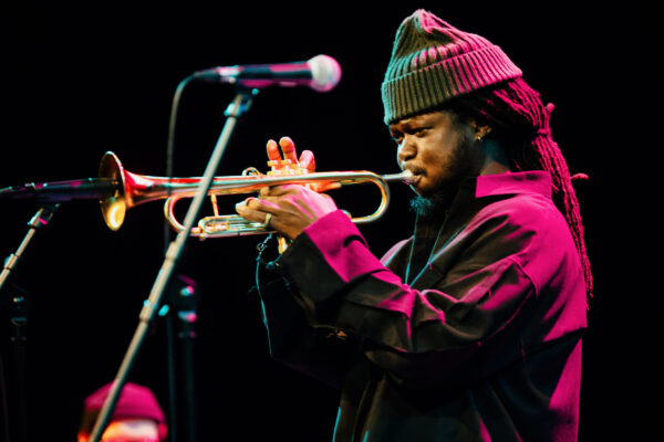 A black man plays the trumpet - he is wearing a hat and a shirt jacket.