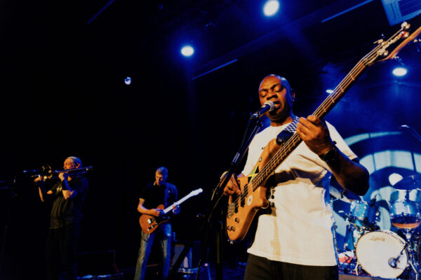 A black man plays bass guitar at the front of a band on stage.