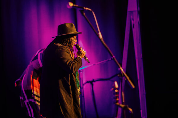 A black woman with dreads and wearing a hat and coat performs on stage with a microphone.