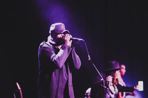 A black man wearing a cap and jacket speaks into a microphone.