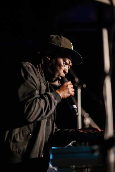 An older black man wearing a cap and jacket speaks into a microphone.