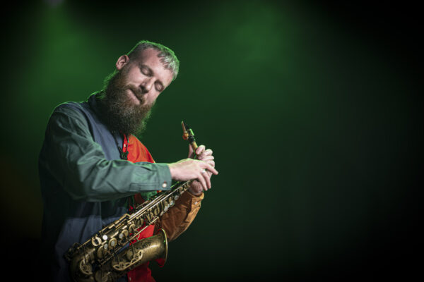 A blonde, white man holding an alto saxophone pauses with his eyes shut. The background is green.