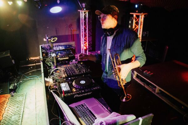 A white man wearing a cap holds a trumpet behind a DJ deck