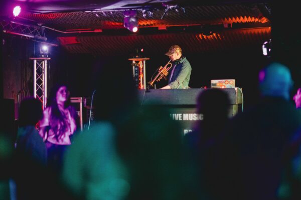 A white man wearing a cap plays the trumpet behind a DJ deck.