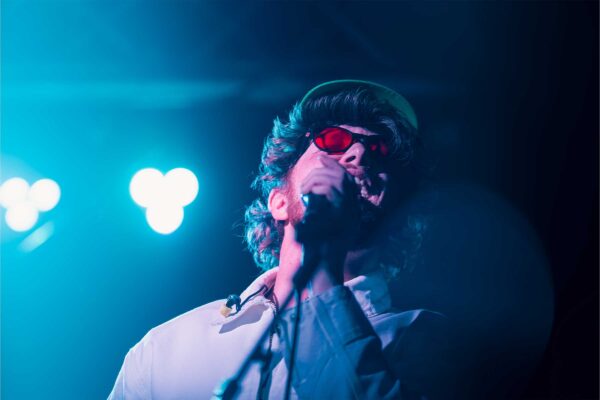 A man lit up in blue sings into a microphone in a cap and red sunglasses