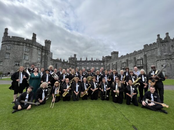 A group of musicians pose with their instruments in front of a stately home, made of grey stone