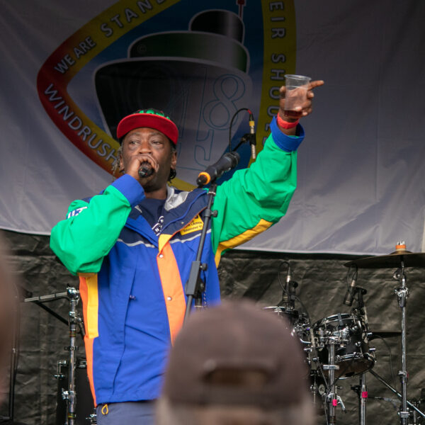 A black man wearing a cap and colourful outfit speaks into a microphone with one hand in the air