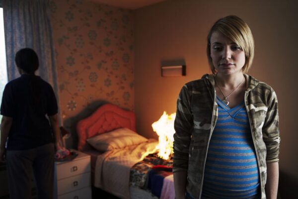 A pregnant woman stands in front of a bed. On the bed, an item of clothing is on fire.