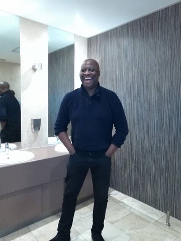 A black man in a black shirt poses, smiling in front of a mirror