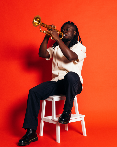 A black man with dreads wearing a white shirt and black trousers plays a trumpet. He is sat on a white ladder and the background is red.