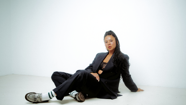 A woman with long, black braids poses in a white room, she is wearing a black suit and silver loafers. There is a soft focus on the camera so the image looks glowy.